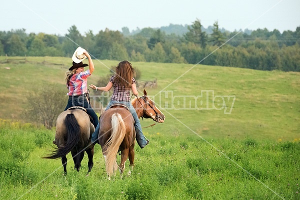 Two young woman horseback riding western goofing around stealing each others cowboy hats.