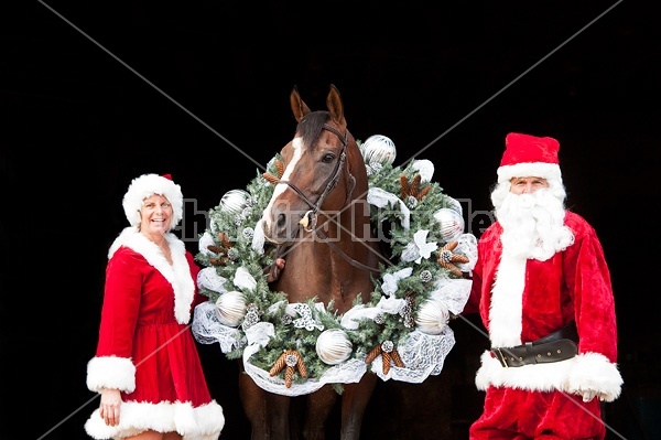 Santa Claus and Mrs Claus standing with a thoroughbred horse with a Christmas wreath over its head.