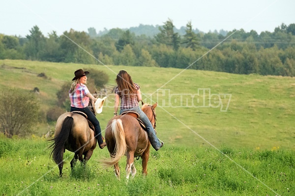 Two young woman horseback riding western goofing around stealing each others cowboy hats.