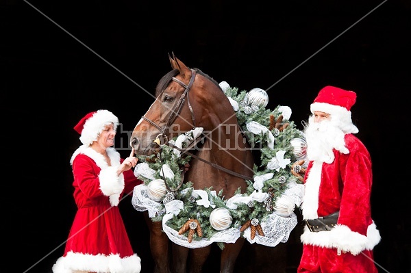 Santa Claus and Mrs Claus standing with a thoroughbred horse with a Christmas wreath over its head.