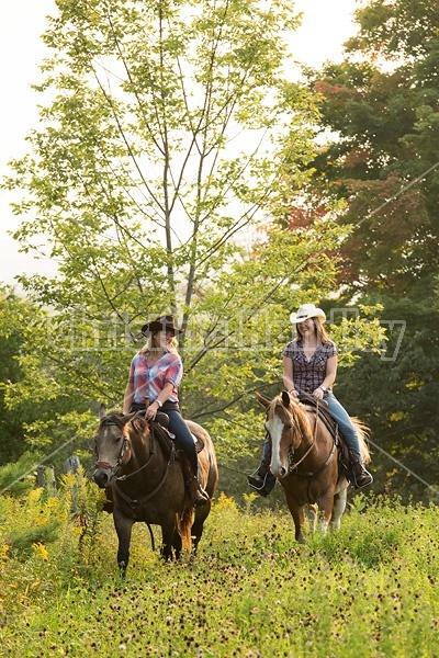 Two young women horseback riding western through summer pasture fields.