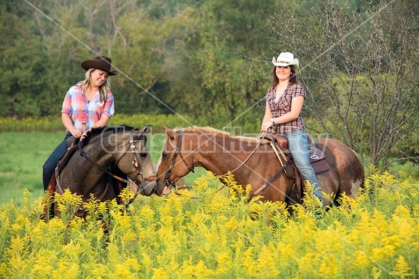 Two young women horseback riding western through summer pasture fields.