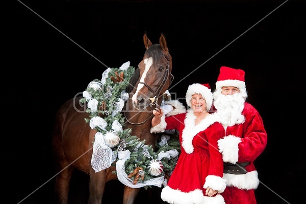 Santa Claus and Mrs Claus standing with a thoroughbred horse with a Christmas wreath over its head.