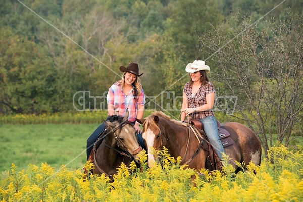 Two young women horseback riding western through summer pasture fields.