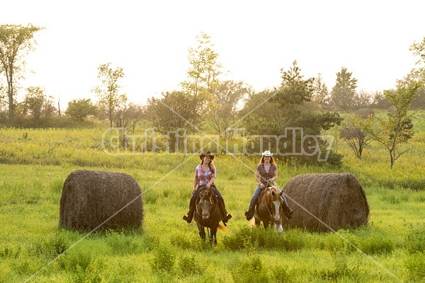 Two young women horseback riding western through summer pasture fields.