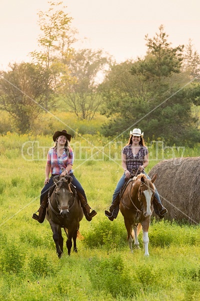 Two young women horseback riding western through summer pasture fields.