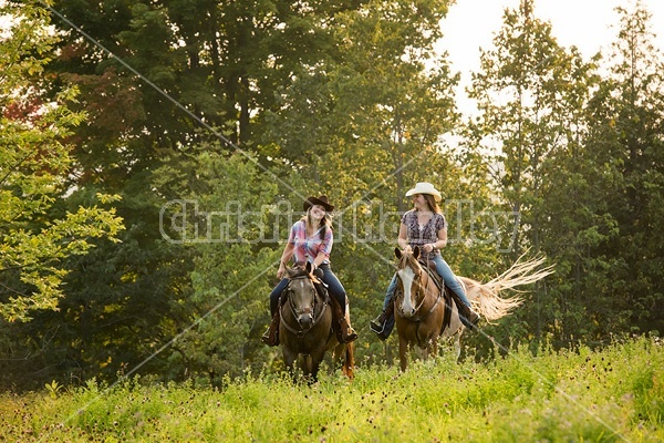 Two young women horseback riding western through summer pasture fields.