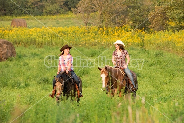 Two young women horseback riding western through summer pasture fields.