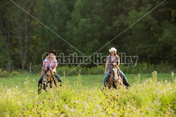 Two young women horseback riding western through summer pasture fields.