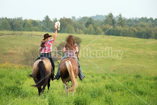 Two young woman horseback riding western goofing around stealing each others cowboy hats.